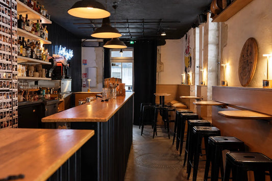 Intérieur de bar étroit avec comptoir en bois, tabourets, étagères de bouteilles et suspensions.